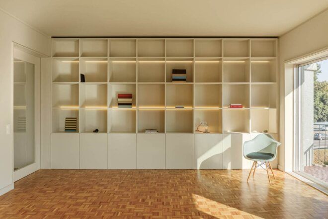 A room with wood floors and white walls with built-in cubbyhole storage cabinets. To the right is a light blue chair next to a large window.