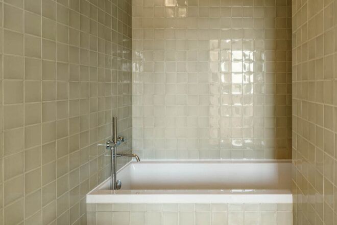 A bathroom with a bathtub surrounded by off-white square tile.