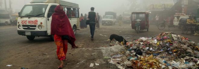 A trash-covered street in Thailand with people and cars going by.
