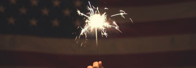 A hand holding a sparkler in front of an American flag.