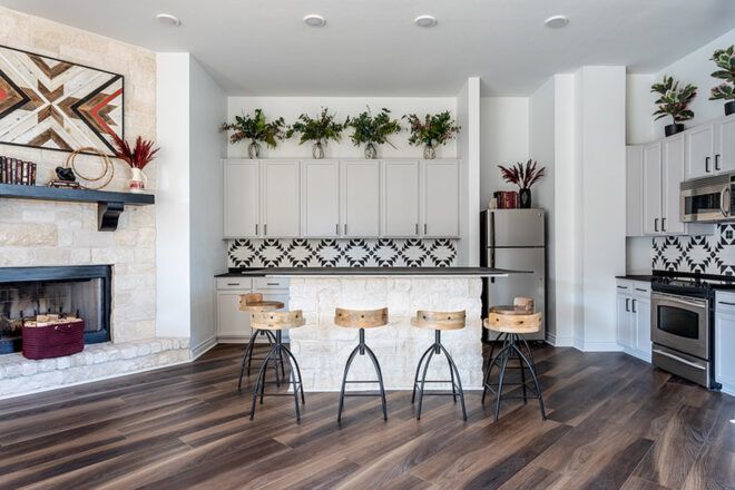 A kitchen with white cabinets and walls and dark wood flooring. There is an island with six wood stools placed around it, a fireplace to the left, and a fridge, stove, oven and microwave to the right.