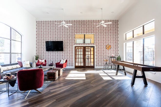 A living room space with white and brick red patterened wall paper on one accent wall, surrounded with two white walls with large windows on the left and right. The room has wood floors and features red furniture and wood tables.