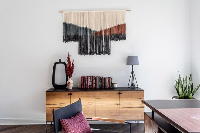 An office space with a chest of drawers against a wall that features a hanging piece of textile art in cream, blue and red colors.