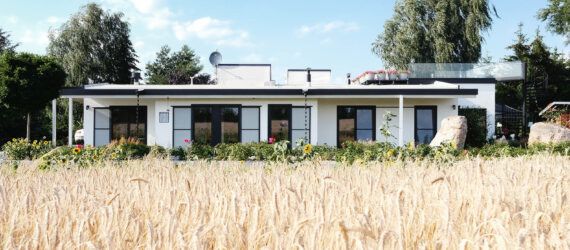 A field of wheat in front of a one-level home with a white and black facade featuring large windows.