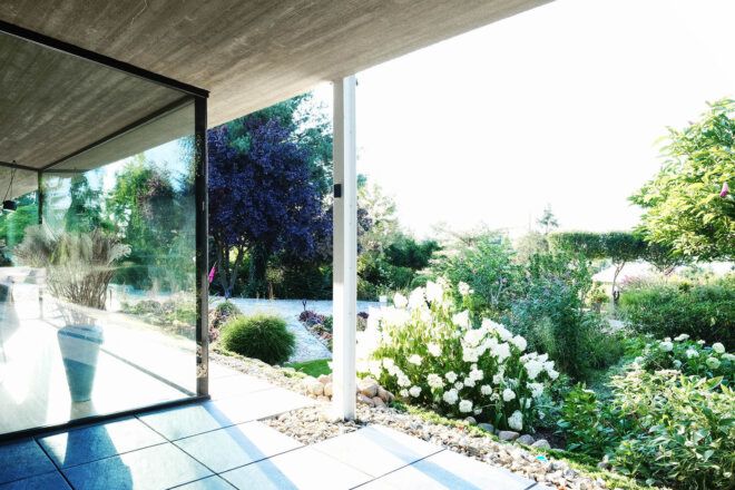 A floor-to-ceiling window to the left of a covered terrace that faces flowers and bushes.