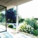 A floor-to-ceiling window to the left of a covered terrace that faces flowers and bushes.
