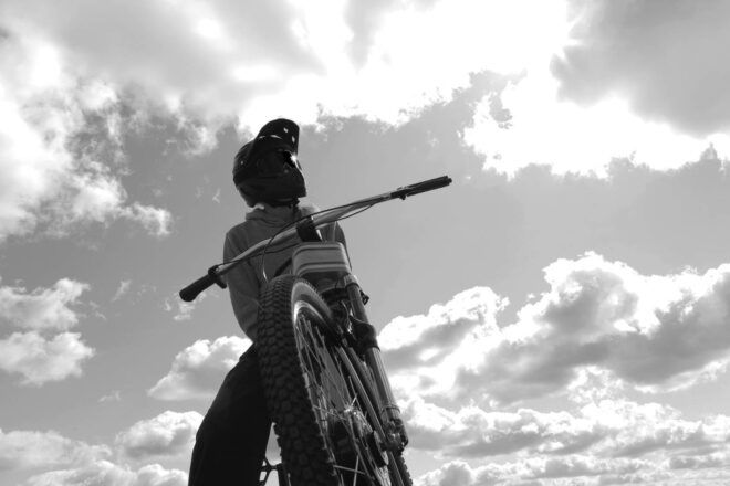 A black-and-white photo of a view of the bike from the front tire, with a person posed atop the bike.