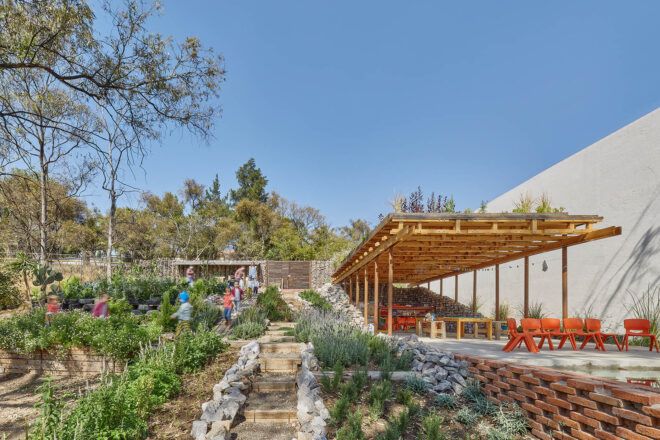 To the left, children walk through a garden. To the right, orange chairs are set up near a wooden pavilion.