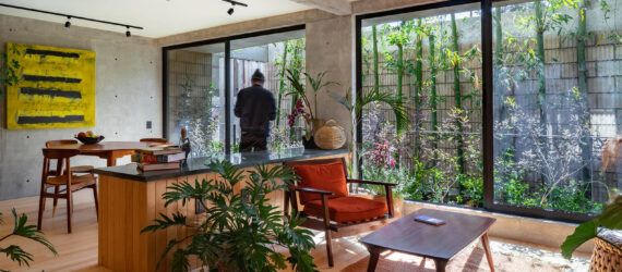 A living room with wood accents, several chairs, two tables and several plants decorating the room. In the background, a person faces a floor-to-ceiling window that looks out on a garden.