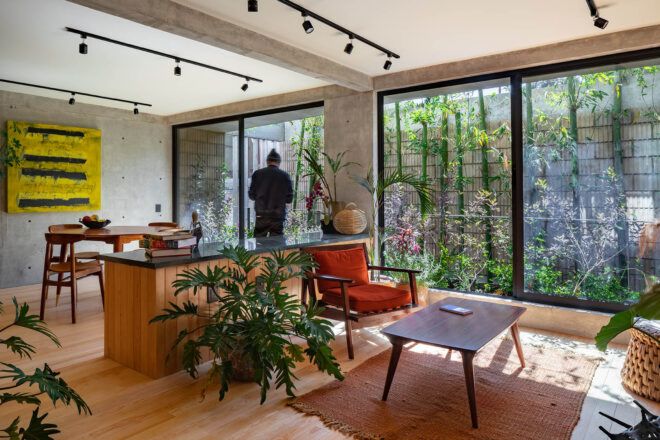 A living room with wood accents, several chairs, two tables and several plants decorating the room. In the background, a person faces a floor-to-ceiling window that looks out on a garden.