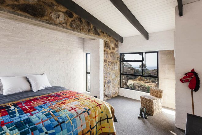 A bedroom with a red, blue and yellow bedspread, stone accents and a window with a dark frame.