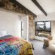 A bedroom with a red, blue and yellow bedspread, stone accents and a window with a dark frame.