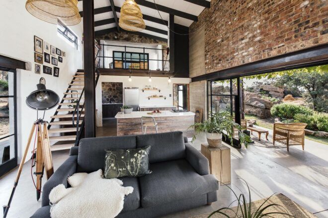 A decorated living room with a gray sofa, white walls and brick and dark wood accents. A staircase in the background leads upstairs. Large sliding glass doors connect the space to the outdoors.