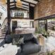 A decorated living room with a gray sofa, white walls and brick and dark wood accents. A staircase in the background leads upstairs. Large sliding glass doors connect the space to the outdoors.