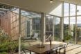 A wood dining table with chairs inside a room surrounded by glass walls that look out onto a green courtyard.