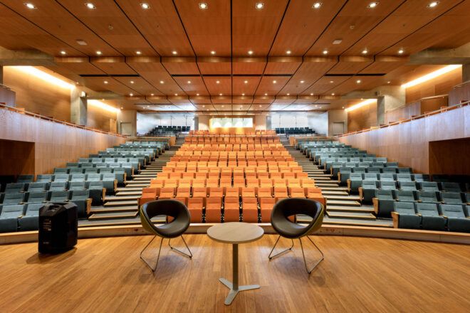 An auditorium with medium-toned wood accents. On stage, two black chairs surround a small circular beige coffee table.