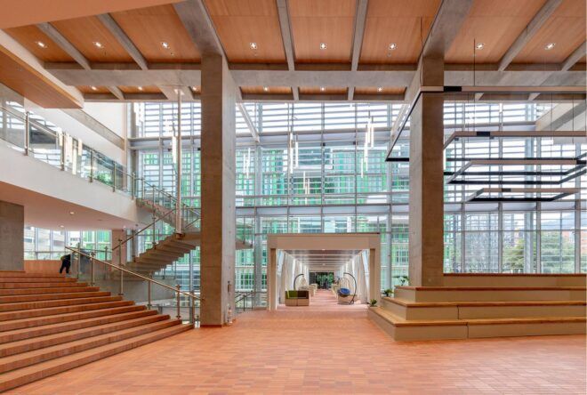 A lobby with a natural orange-toned material palette. Glass walls frame the room, which features a staircase to upper levels.
