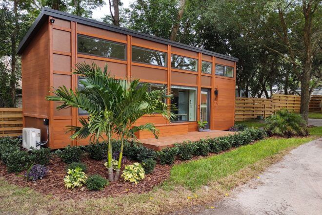 A rectangular tiny home on an elevated porch, with orange-toned wood siding. The home is surrounded by plants.