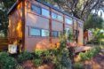 A rectangular tiny home on an elevated porch, with orange-toned wood siding. The home is surrounded by plants.