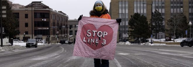 A woman holds a pink Stop Line 3 sign in the middle of the street.