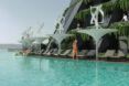 A pool area lined with white fixtures topped with helixes. In the background, people stand on their balconies on the hotel building.
