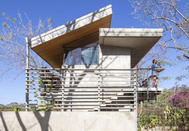 A white house with an assymetrical roof. A parent carries a baby on the outdoor staircase.
