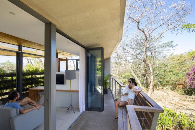 To the left, a person sits on a gray armchair inside. To the right, large glass doors open onto a balcony where a person sits with a baby.