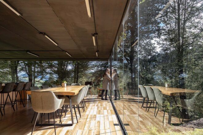 A dining room with light-toned wood floors and a dining table with chairs. The room is surrounded by floor-to-ceiling glass walls, and a person on the right looks out through them.