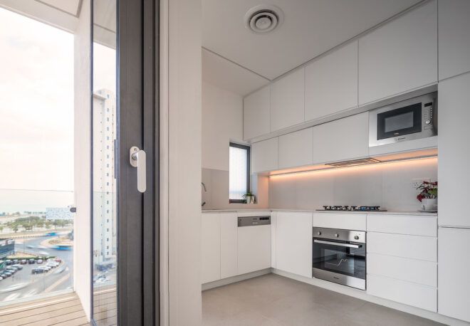 To the left, a glass door leading to an outdoor balcony. To the right, a kitchen in all white.