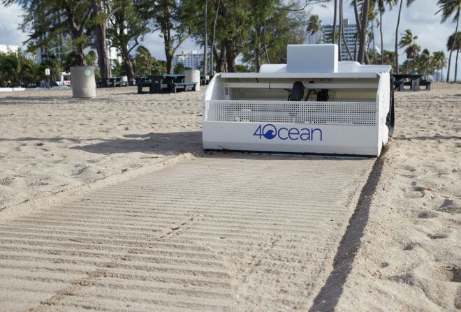 A small white robot with wheels and a 4ocean logo on it sweeps a beach for plastic waste.