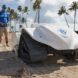 A person in shorts, a blue shirt and hat operates a small white robot on wheels via remote control.
