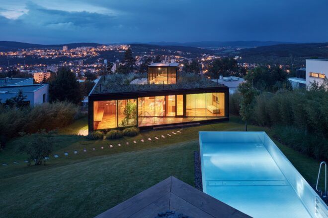 A backyard with a rectangular pool that looks onto the back of a rectangular house with a green roof. In the background is a city lit up in the evening.