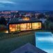 A backyard with a rectangular pool that looks onto the back of a rectangular house with a green roof. In the background is a city lit up in the evening.