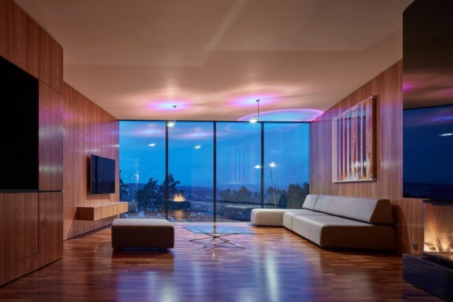 A living room with a gray sofa, TV, coffee table and ottoman. The parallel wall features floor-to-ceiling windows.