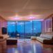 A living room with a gray sofa, TV, coffee table and ottoman. The parallel wall features floor-to-ceiling windows.