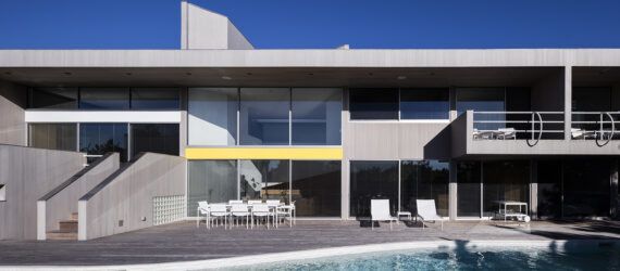 A pool connects to a deck with white patio chairs. This sits in front of a two-level house with floor-to-ceiling windows.