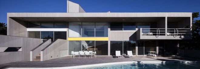 A pool connects to a deck with white patio chairs. This sits in front of a two-level house with floor-to-ceiling windows.