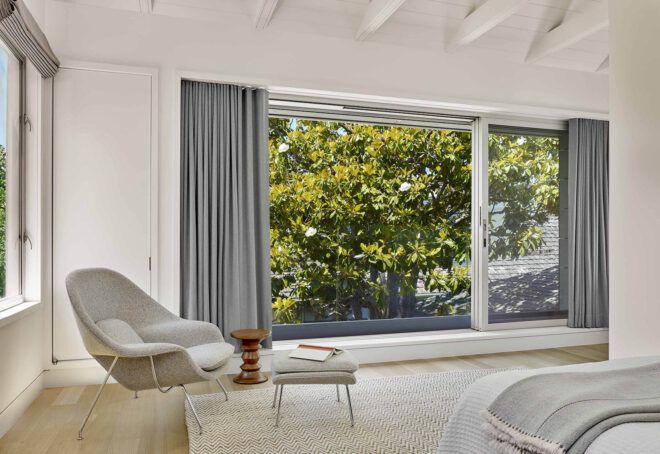 A white bedroom with a chair next to a large window framed by gray curtains.