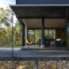 A single-level cabin with a roof over a gray patio that borders a forest.
