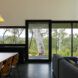 An interior living space where a dining area connects to a living room, both bordered by a wall of glass overlooking a forest.
