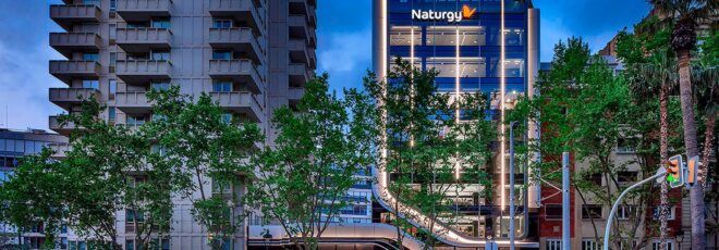 A nighttime street lined with trees that partially obscure the facade of a glass business tower with a logo reading "Naturgy."