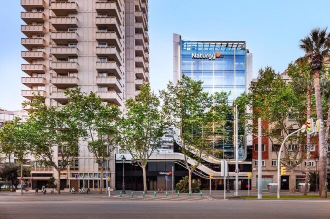 A daytime street lined with trees that partially obscure the facade of a glass business tower with a logo reading "Naturgy."