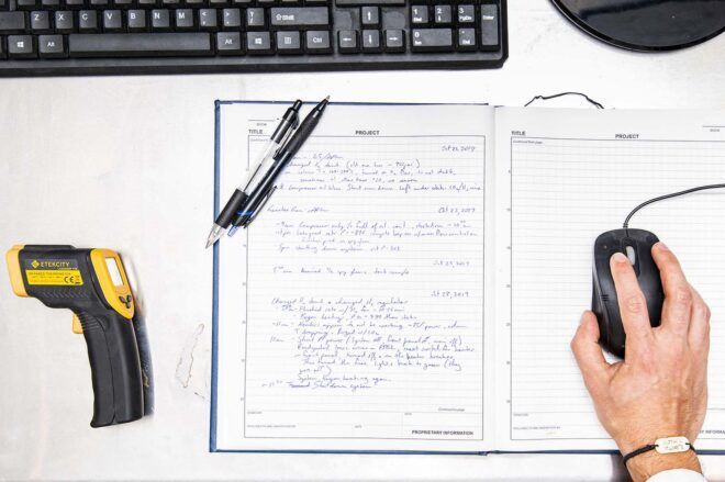 A work desk with a yellow and black tool, a notebook and a hand holding a computer mouse.