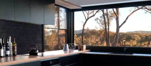 A kitchen with black cabinets with wood countertops. A large window above the sink overlooks a forest.