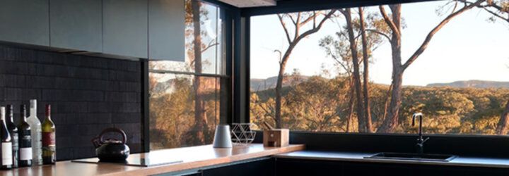 A kitchen with black cabinets with wood countertops. A large window above the sink overlooks a forest.