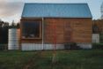 The front of a simple wood house with a steel water tank to the left and a small red tree in front of the house.