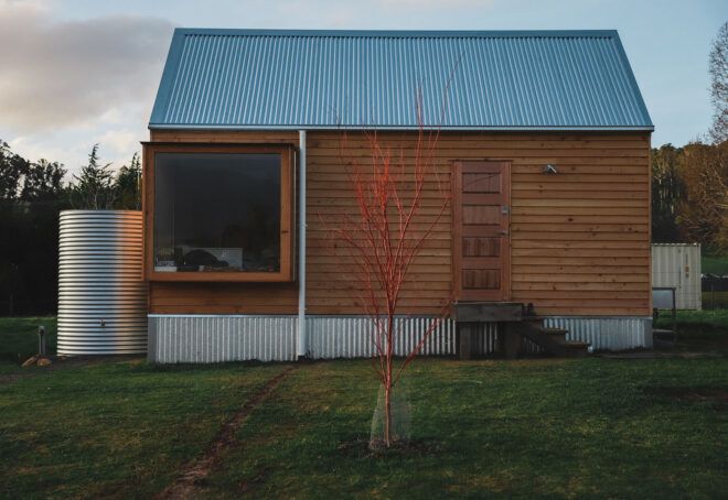The front of a simple wood house with a steel water tank to the left and a small red tree in front of the house.