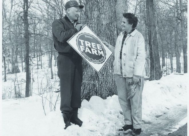 A black and white photo of a couple standing in the snow, with the person on the left holding a sign that says, "tree farm."