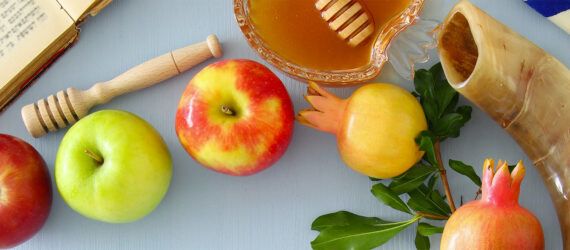 A spread of apples, honey, pomegranates and a horn on a blue-gray table.