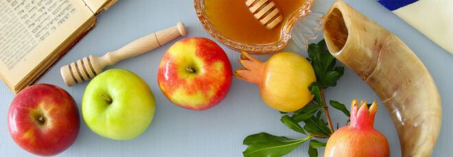 A spread of apples, honey, pomegranates and a horn on a blue-gray table.
